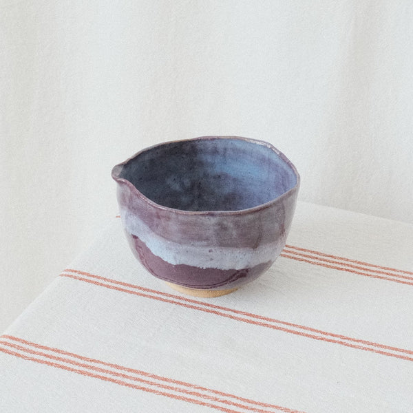 A small handmade ceramic bowl with a rustic, hand-painted finish in shades of purple, blue, and white, resting on a cream-colored tablecloth with thin red stripes against a soft white fabric backdrop.